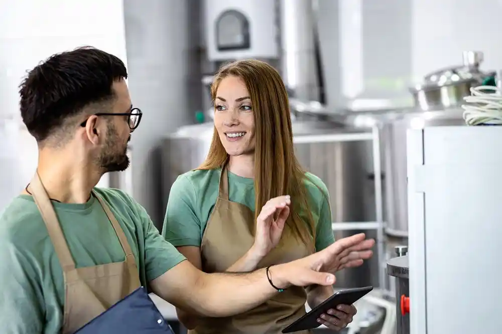 Bir mutfakta önlüklü iki çalışan konuşurken biri tablet tutuyor